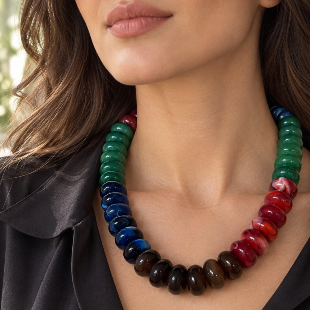 A close-up of a woman wearing a chunky beaded necklace made of glossy, polished oval beads in a gradient of colors from green and blue to red and dark brown.