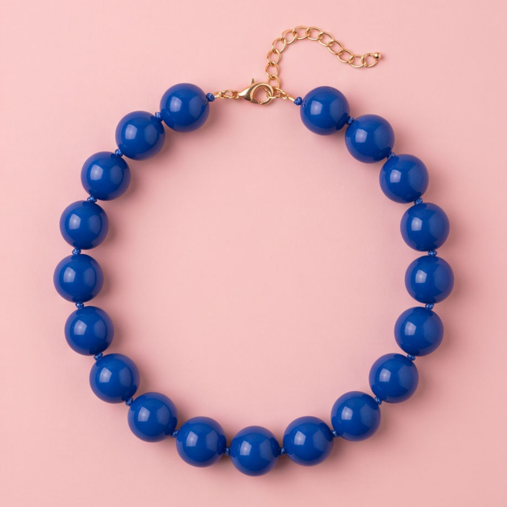 A beaded necklace with glossy, large round blue beads and a gold-tone chain extender clasp, displayed on a solid light pink background.