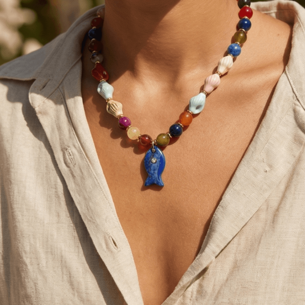 Colorful beaded necklace with a blue fish pendant worn by a person in a beige shirt.