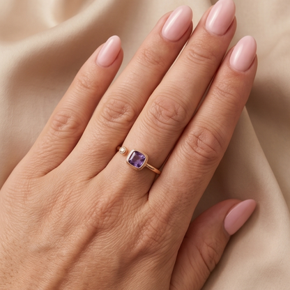 Hand wearing a gold ring with a purple gemstone on a beige fabric background