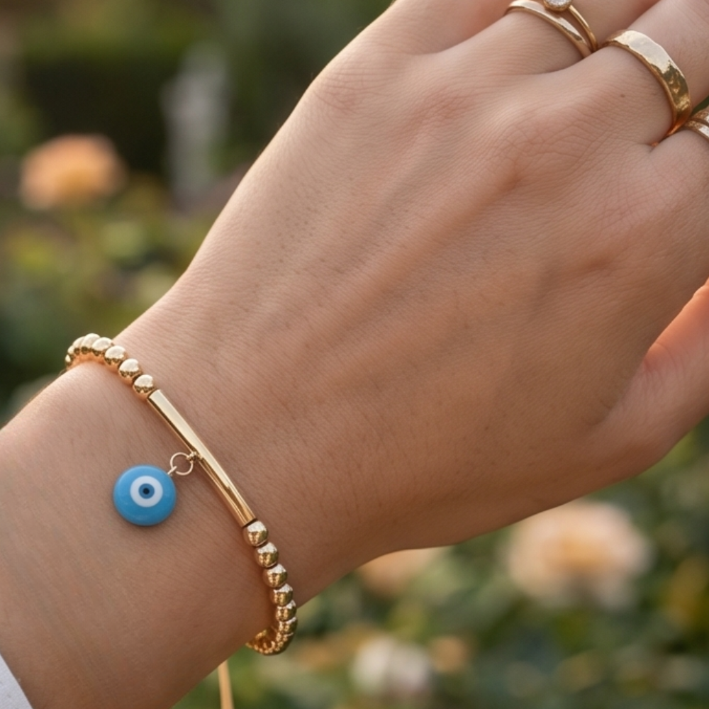 Gold bracelet with a blue evil eye charm on a wrist against a blurred natural background