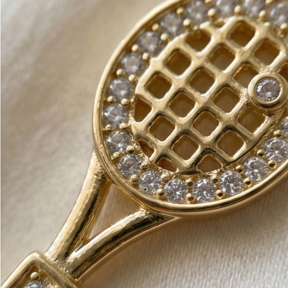 Gold tennis racket-shaped object with diamond-studded strings on a beige background
