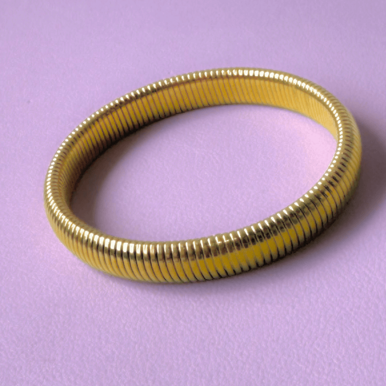 A close-up image of an elegant gold-plated coil bangle, showcasing its unique coiled design and polished finish, placed on a rustic white wooden surface with a string of pearls.