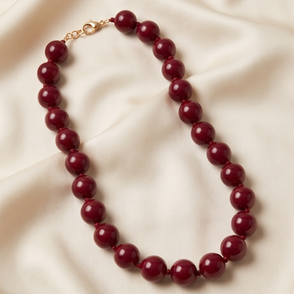 A beaded necklace made of glossy, deep burgundy red round beads with a gold-tone clasp, displayed on a draped cream satin fabric background.
