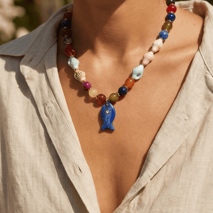 Colorful beaded necklace with a blue fish pendant worn by a person in a beige shirt.