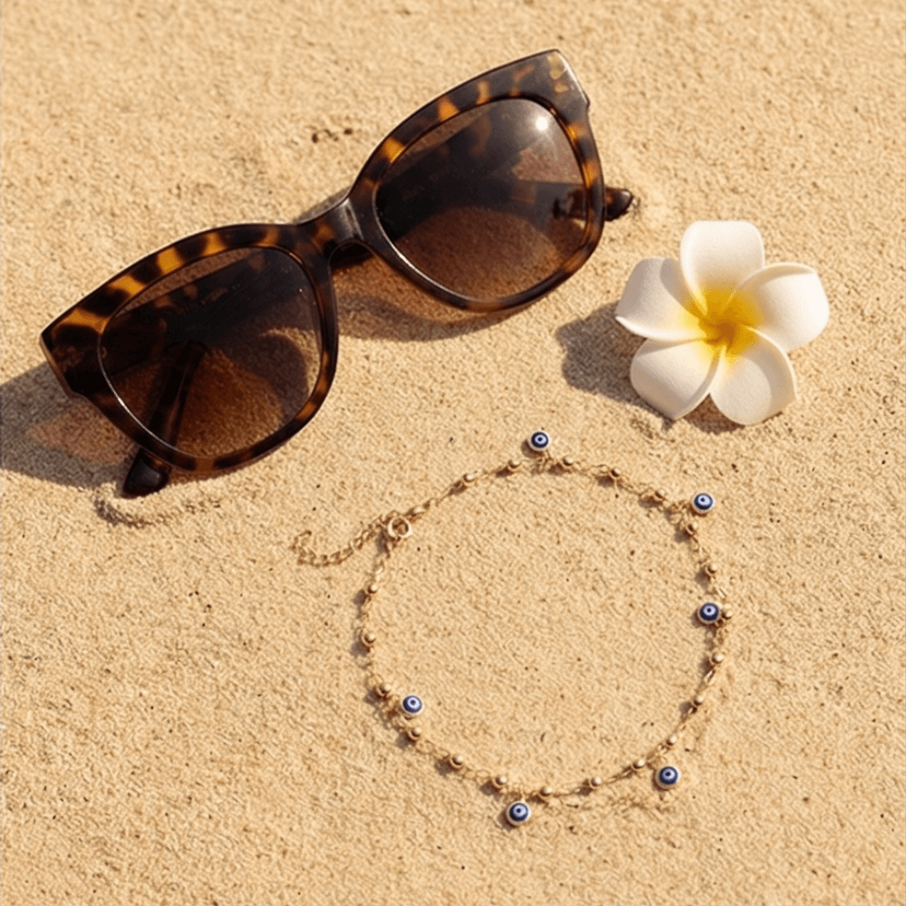 Gold evil eye anklet bracelet for women styled on sand with sunglasses, delicate summer ankle jewelry.
