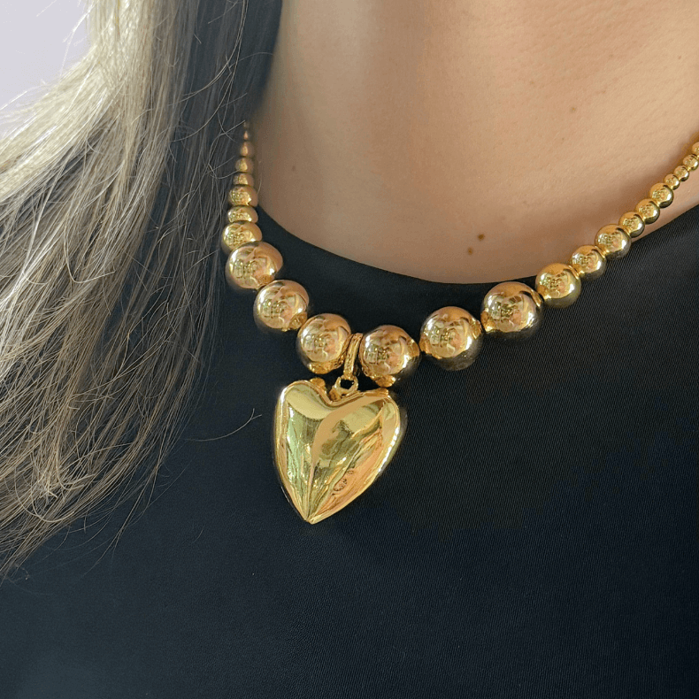 This image shows a gold necklace with large, round beads and a chunky, polished heart-shaped pendant in the center. The beads vary in size, with larger beads toward the front and smaller ones toward the back. The necklace has a glossy finish, creating reflections on the gold surface, and is worn against a dark-colored top.