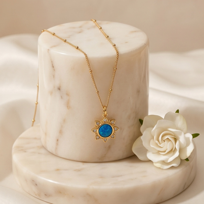 Gold blue opal sun pendant necklace displayed on marble stand with white rose
