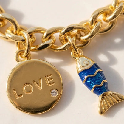 Gold chain charm bracelet with love pendant and blue fish charm displayed on jewelry stand by Julie Saldivia Jewelry