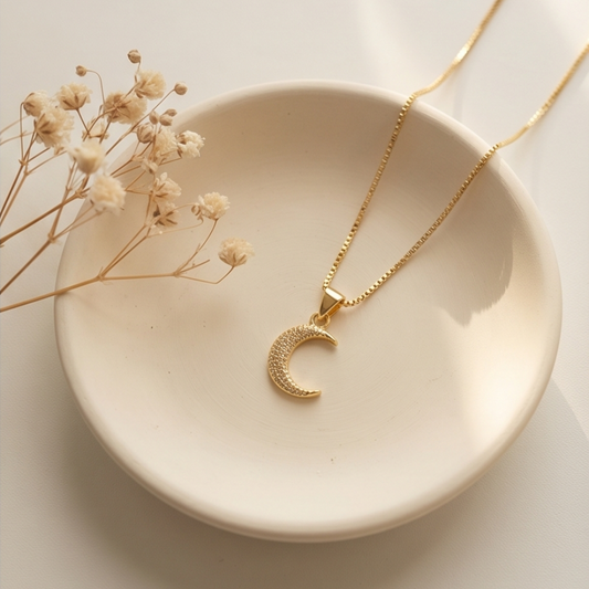 Gold necklace with a crescent moon pendant on a beige dish with dried flowers.