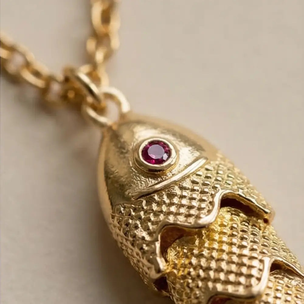 Gold fish-shaped pendant with a red gemstone on a light background