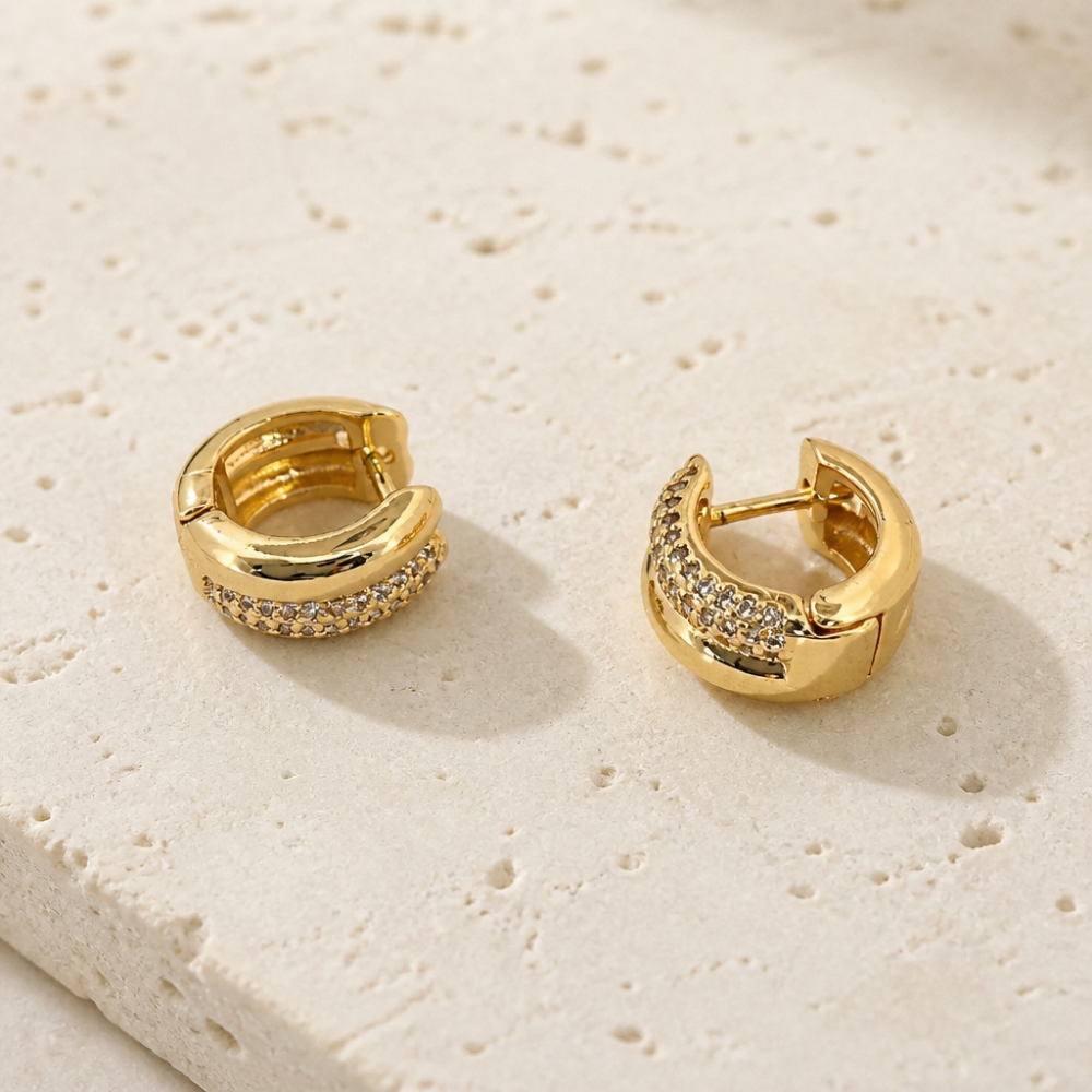 A pair of gold huggie hoop earrings with pavé-set clear stones, displayed on a textured beige stone surface.