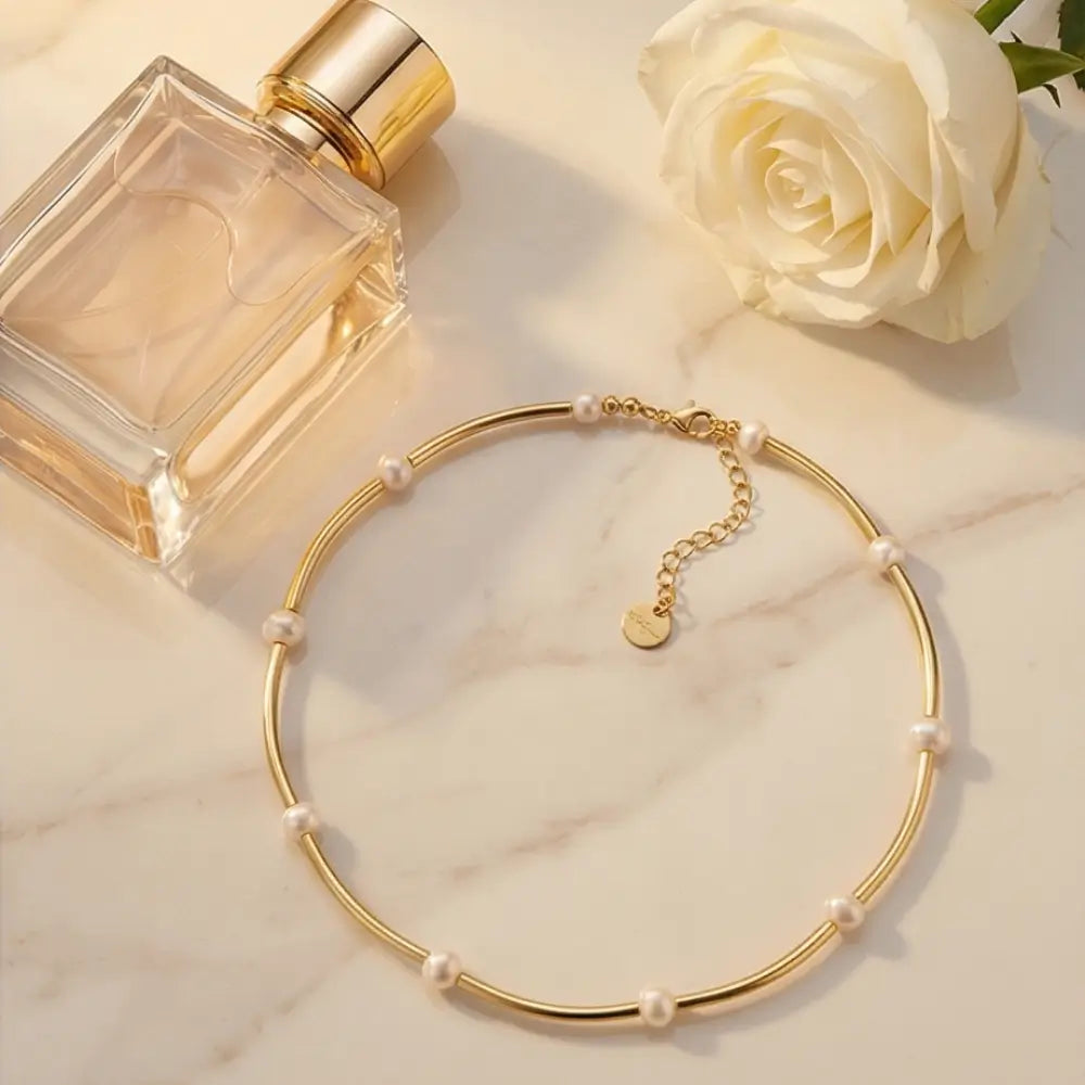 Gold necklace with pearls next to a perfume bottle and white rose on a marble surface