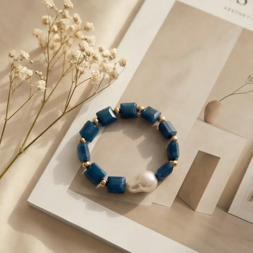 Blue beaded bracelet with a pearl on a light wooden surface with dried flowers.