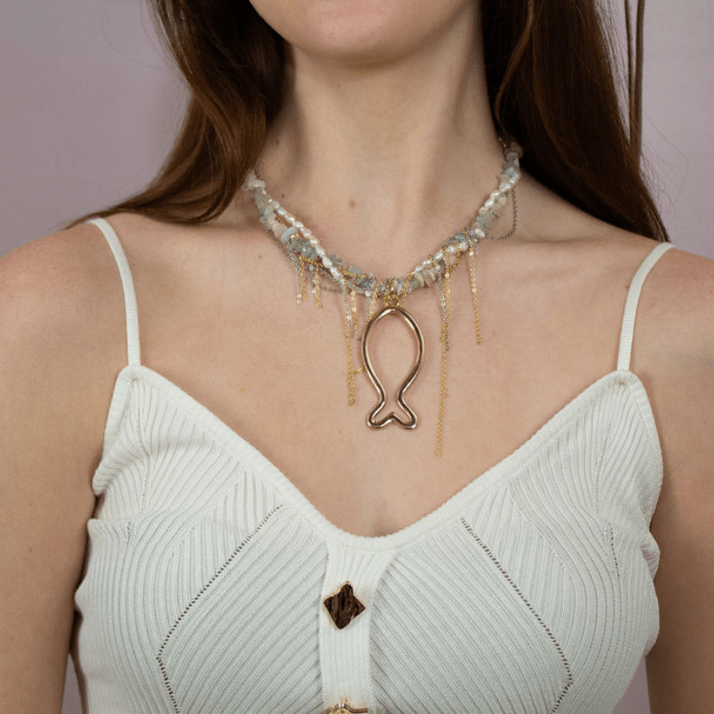 A woman in a white top and cowboy hat holding a handmade necklace featuring a fish design, adorned with pearls and natural stones, with gold chains.