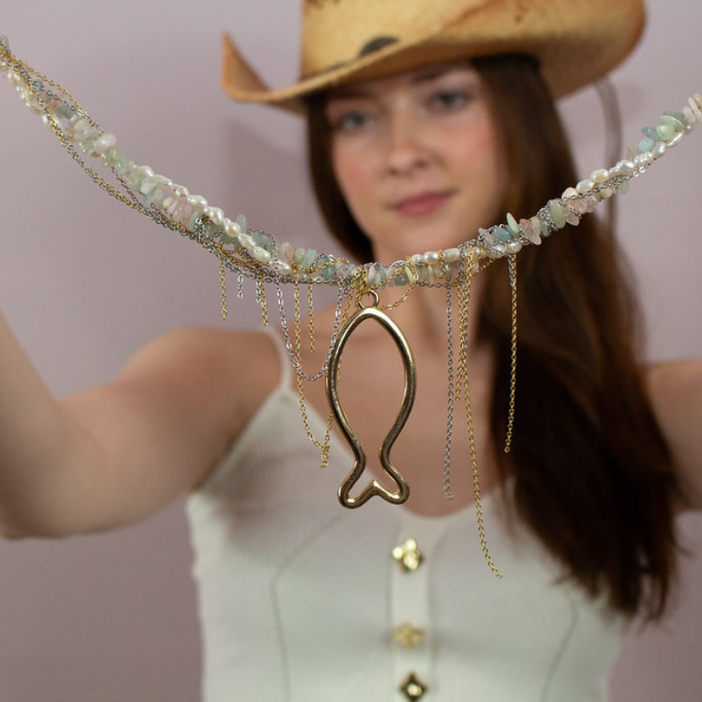 A woman in a white top and cowboy hat holding a handmade necklace featuring a fish design, adorned with pearls and natural stones, with gold chains.