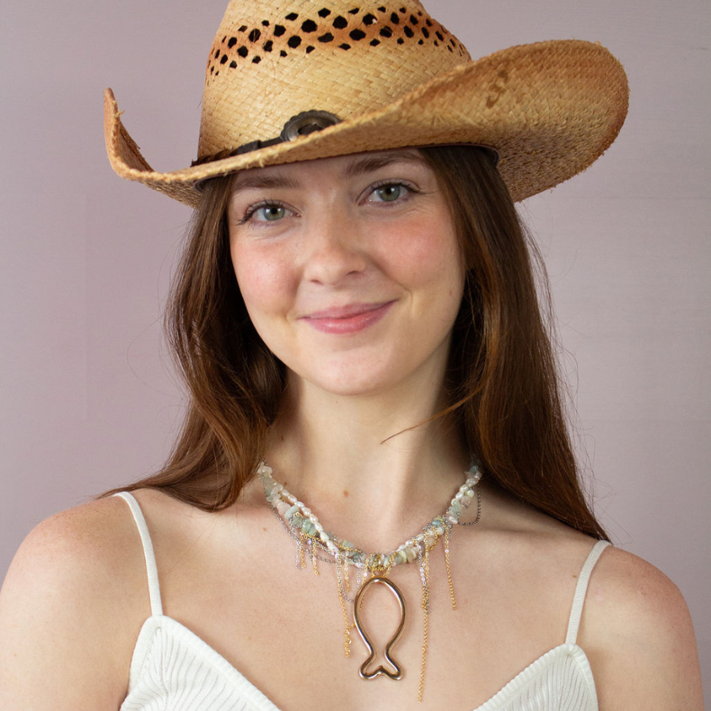 A woman in a white top and cowboy hat holding a handmade necklace featuring a fish design, adorned with pearls and natural stones, with gold chains.