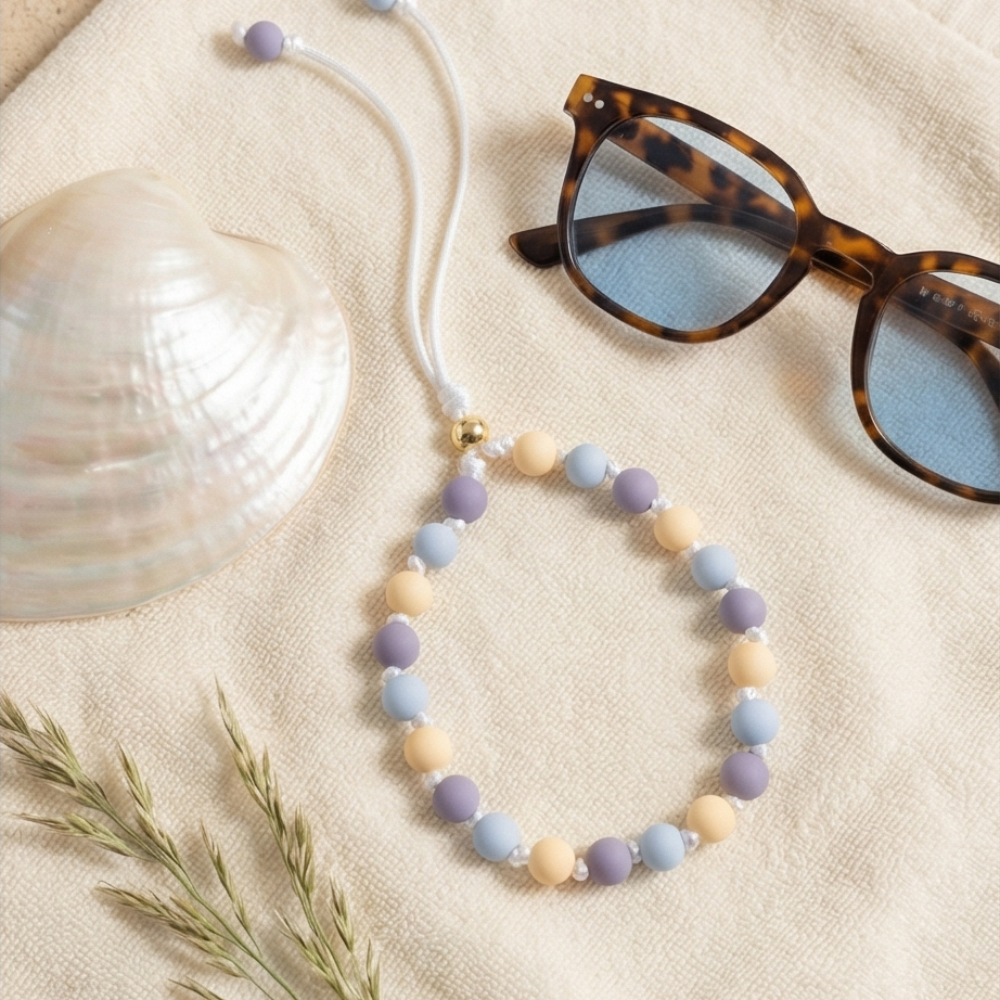 Beaded necklace with pastel colors on a sandy surface with sunglasses and a shell.