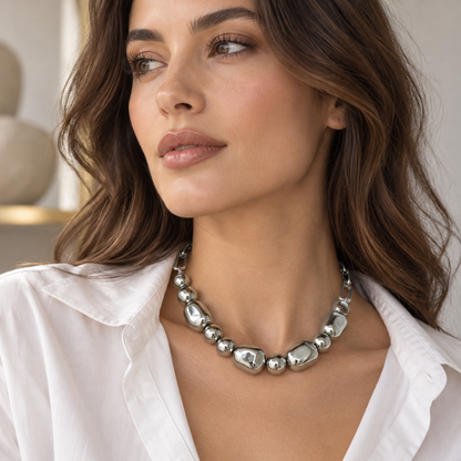 A close-up of a woman wearing a chunky silver statement necklace made of irregular polished beads, paired with a white collared shirt.