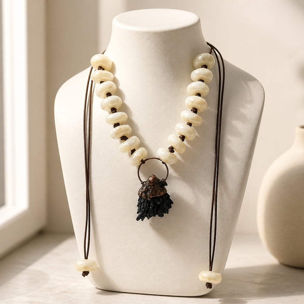 A bohemian-style beaded necklace with cream-colored disc beads, dark brown cords, and a dark brown textured tassel pendant, displayed on a white ceramic bust.