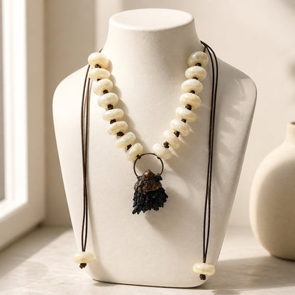 A bohemian-style beaded necklace with cream-colored disc beads, dark brown cords, and a dark brown textured tassel pendant, displayed on a white ceramic bust.