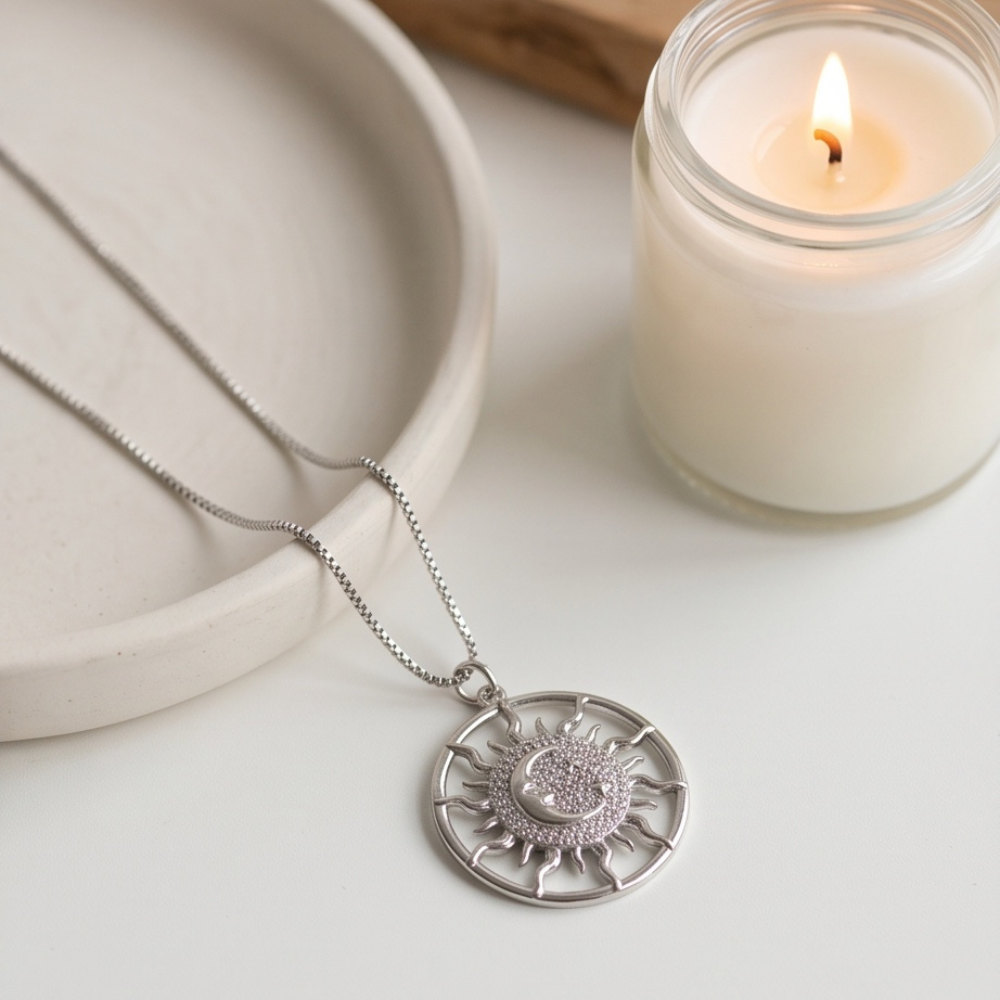 Silver sun and moon pendant necklace displayed beside a white candle and ceramic dish