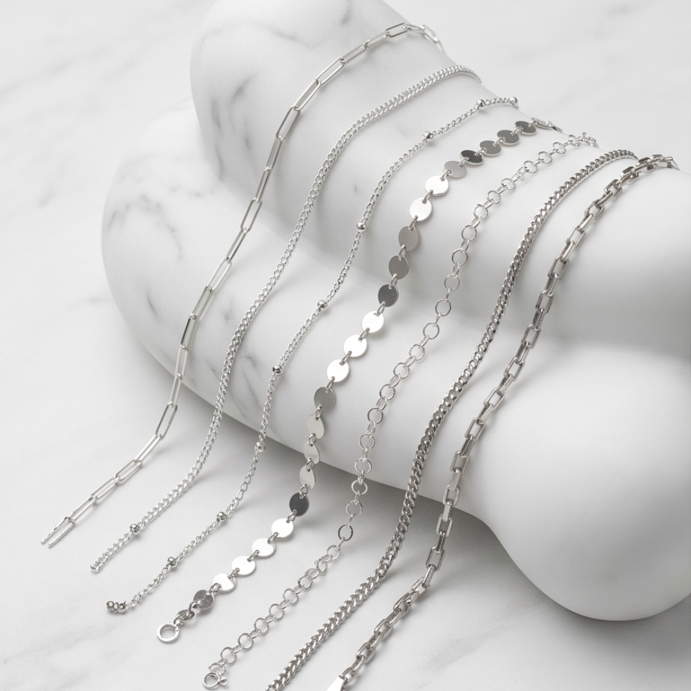 Selection of silver permanent jewelry chains including paperclip, disc, beaded, and classic link styles.