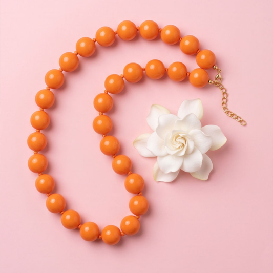 An orange beaded necklace with a gold-tone clasp, arranged on a light pink background next to a white artificial flower.