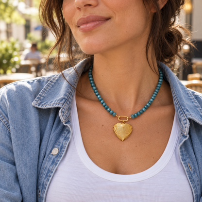 Woman wearing the 18-inch Tidal Heart aventurine necklace with an 18k gold-plated ridged heart pendant, styled with a white top and denim shirt at an outdoor café.