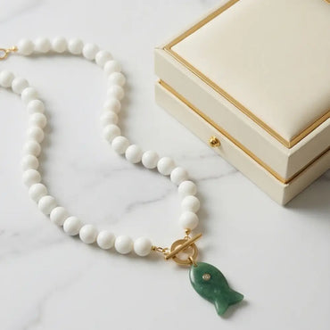 White beaded necklace with green fish pendant displayed next to jewelry box minimalist luxury styling